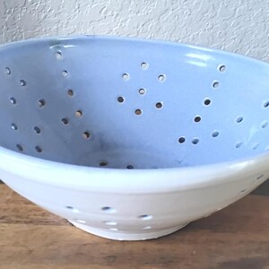 Puede incluir: Un colador de cerámica con interior azul claro y exterior blanco. El cuenco tiene numerosos agujeros pequeños para escurrir y dos asas pequeñas. El colador está sobre una superficie de madera.