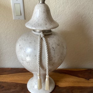 May include: A large, white ceramic vase with a marbled pattern and a silver lid. The vase has a rope-like trim with tassels hanging from the bottom.