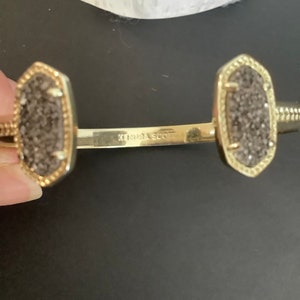May include: A gold Kendra Scott cuff bracelet with two grey stone charms. The bracelet is open and has a hinge.