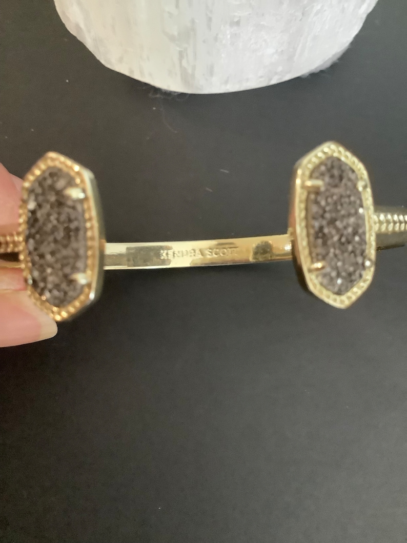 May include: A gold Kendra Scott cuff bracelet with two grey stone charms. The bracelet is open and has a hinge.