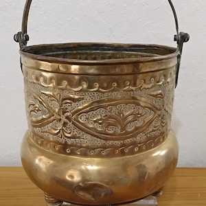 May include: A round brass bucket featuring an embossed floral pattern around the center, a smooth base, and a curved metal handle attached to the sides. The vessel stands on small feet and displays a polished metallic finish with intricate detailing.