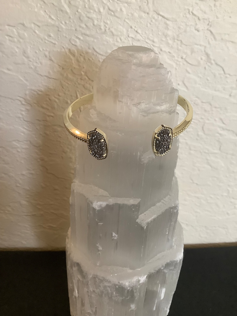 May include: A gold bangle bracelet with two grey stone accents. The bracelet is displayed on a white crystal.