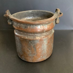 May include: A vintage copper pot with two handles. The pot has a worn, aged patina and is slightly dented.