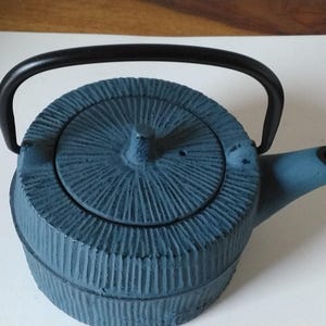 Puede incluir: Una tetera de hierro fundido azul con un patrón texturizado de ranuras radiales en la tapa y el cuerpo. La tetera incluye un asa negra arqueada y un pico azul a juego con una abertura interior negra, con un acabado mate en todo el objeto.