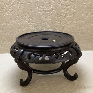 May include: A dark brown wooden display stand featuring a circular top with a central hole and a small yellow sticker. The base is supported by four curved legs with intricate cut-out patterns along the sides of the circular frame.