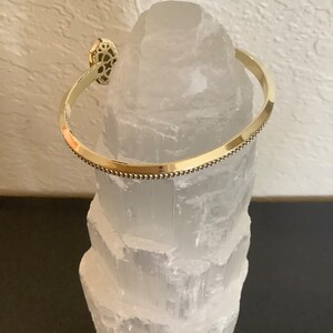 May include: A gold-toned bangle bracelet with a decorative design on the front. The bracelet is resting on a white selenite crystal.