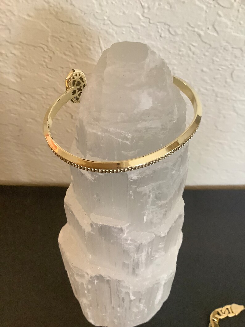 May include: A gold-toned bangle bracelet with a decorative design on the front. The bracelet is resting on a white selenite crystal.
