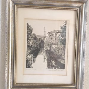 May include: Framed black and white print of a canal scene with buildings and reflections in the water. The print is in a silver and gold ornate frame. The artwork is signed and numbered.