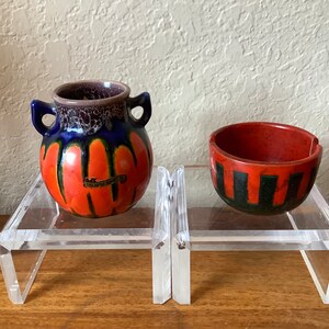 May include: Two small ceramic pieces, a red and black bowl and a blue, orange, and black vase with a label that reads "Deutsche Handarbeit". The pieces are displayed on a clear acrylic stand.