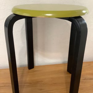 May include: A small, round stool with a lime green seat and black legs. The legs are curved and angled, providing a sturdy base. The stool is made of wood and has a simple, minimalist design.