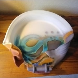 May include: A white ceramic bowl with a unique, abstract design. The bowl has a wavy, folded edge and features a blue, green, and brown glaze.