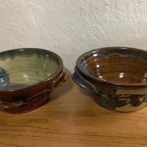 May include: Two round ceramic bowls with small side handles. The left bowl features a light green interior with a dark brown exterior. The right bowl has a deep brown interior and a mottled light green and dark brown exterior with a glossy finish.