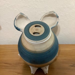 Könnte beinhalten: Ein rundes Sparschwein aus Keramik mit horizontalen Streifen in Blau, Creme und Beige. Die Oberseite hat einen Münzschlitz und zwei große, aufrechte Ohren mit bläulichen Rändern. Der Boden steht auf vier kleinen, weißen, spitzen Füßen.