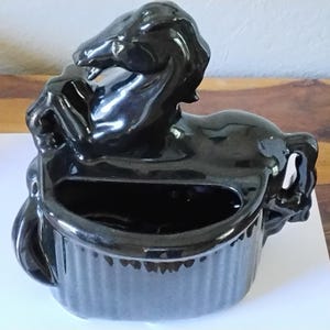 Op de afbeelding: Een zwarte keramische paardvormige plantenbak. Het paard staat op, met een rechthoekige opening om in te planten. De plantenbak heeft een glanzende afwerking en staat op een wit oppervlak.