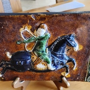 May include: A ceramic tile with a green and brown border depicting a rider on horseback. The rider is wearing a green and white outfit and is holding a bow and arrow. The horse is black and white.