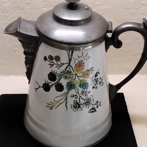 May include: White ceramic coffee pot with a floral design featuring black berries and colorful leaves. The pot has a silver lid, spout, and handle. A decorative piece for the kitchen or dining room.
