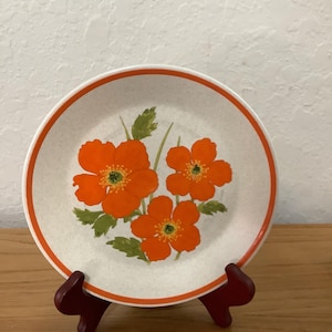 May include: A round, white plate with an orange floral design and a thin orange border. The flowers are bright orange with yellow centers and green leaves. The plate is displayed on a small, dark red stand.