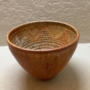 May include: A round ceramic bowl with a speckled, mottled brown and tan glaze. The interior features a series of horizontal ridges along the sides and a subtle, repeating triangular pattern near the rim, showcasing a textured, earthy finish.