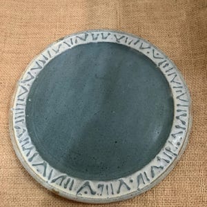 May include: Handmade ceramic plate with a teal center and a textured, off-white rim. The rim features a repeating pattern of stylized markings. The plate's design suggests a rustic or artisanal aesthetic, suitable for serving food or as a decorative piece.