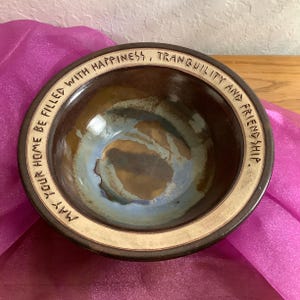 May include: A round ceramic bowl with a dark brown rim and a light brown and blue glazed interior. The rim features the embossed text: MAY YOUR HOME BE FILLED WITH HAPPINESS, TRANQUILITY AND FRIENDSHIP. The bowl has a smooth, glossy finish.
