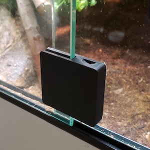 May include: A black, rectangular terrarium door lock. The lock is made of a solid, matte material and is designed to secure a glass door. The lock is attached to the glass door and frame.