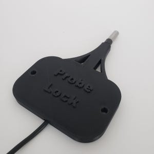 May include: A black plastic "Probe Lock" with a metal probe tip. The device has the words "Probe Lock" embossed on the front. A black wire extends from the base of the device. Two holes are visible on the base.