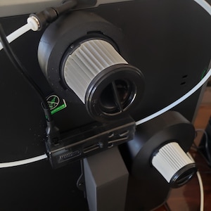 May include: Close-up of a black machine component with two cylindrical air filters. The filters are light gray with black rims. A black rectangular component is attached to the machine. White tubing and a black cable are also visible.