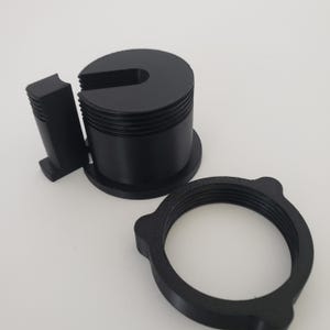 May include: A black cylindrical component with a U-shaped cutout and external threading, accompanied by a separate black rectangular piece with ribbed edges and a black circular ring featuring internal threading and three small protruding tabs.