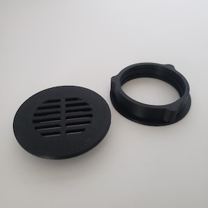 May include: Black, circular drain cover with horizontal slots, next to a black, circular threaded ring. The drain cover is flat and has a series of parallel slots for water drainage. The threaded ring has a raised edge and is designed to secure the drain cover.