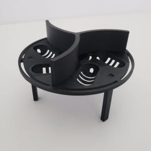May include: A black, three-legged, circular object with a divided top. The top has three curved dividers and cut-out patterns. The object appears to be made of a matte black material.
