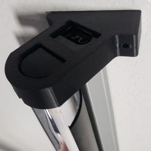 May include: A black, 3D-printed bracket, mounted on a white wall, designed to hold a cylindrical object. The bracket has a curved, recessed area and a hole for mounting. The cylindrical object is silver and has the number "34" printed on it.