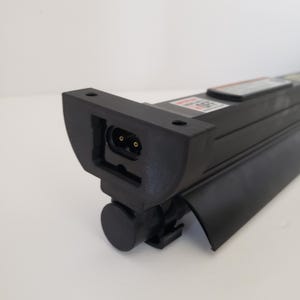 May include: A black electronic component featuring a rectangular housing with a two-pin power connector port on the end. The device includes a cylindrical cap, a curved black panel, and a warning label with the text OFF visible on the top surface.
