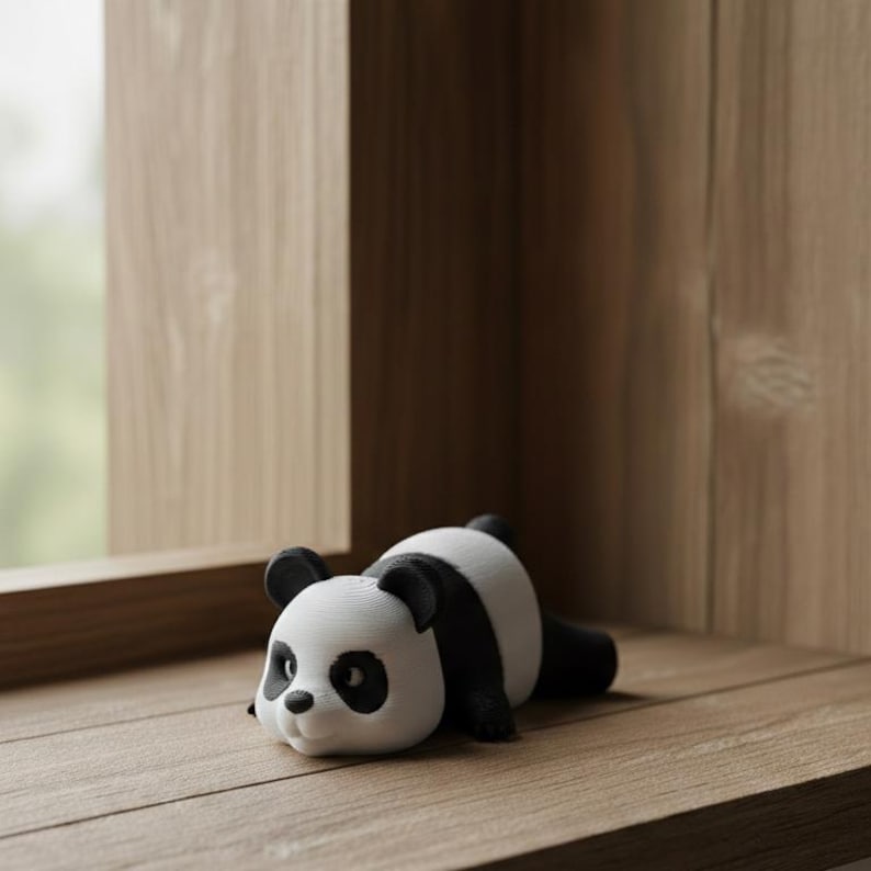 May include: A small, white and black panda figurine resting on a wooden surface. The panda has black ears, eyes, and limbs, with a white face and body. The figurine is lying down, appearing relaxed. The background includes a window and wooden paneling.