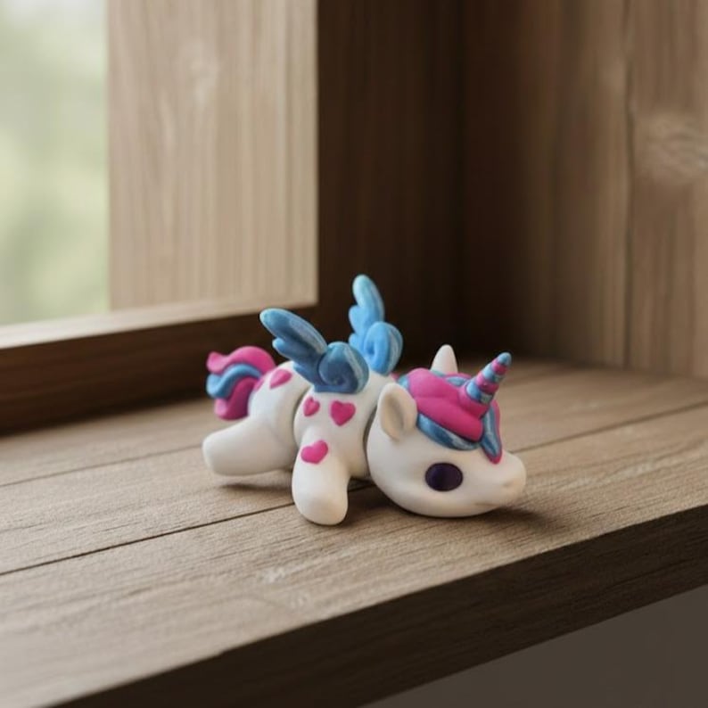 May include: A white unicorn figurine with pink hearts, a pink and blue horn, and blue wings. The toy is lying down on a wooden surface. The unicorn has purple eyes and a pink and blue mane.