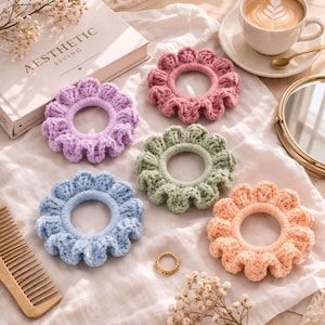 May include: Five handmade, flower-shaped scrunchies in pastel colors: lavender, pink, green, blue, and peach. The scrunchies are made of a textured, crocheted material. A book with the text "AESTHETIC LIVING" is in the background.