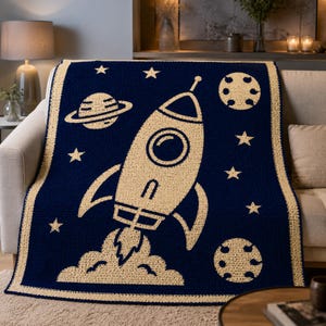 May include: A navy blue crochet blanket featuring a large cream-colored rocket ship launching into space. The design includes a planet with rings, two cratered moons, and several stars scattered across the dark background, finished with a cream border.