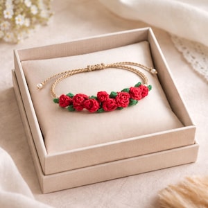 Könnte beinhalten: Ein handgefertigtes Armband mit einer Reihe roter gehäkelter Rosen und grünen Blättern. Das Armband ist auf einem beigen Kordelband und in einer beigen Geschenkbox präsentiert. Die Rosen sind der Blickfang und verleihen einen floralen Akzent.