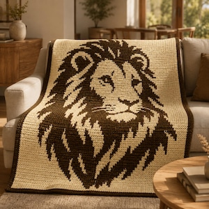 May include: A beige and brown crocheted blanket with a lion's head design. The blanket is draped over a light-coloured sofa. The lion's mane and facial features are in dark brown, contrasting with the lighter background. The blanket is a cozy home decor item.