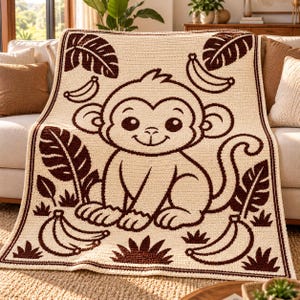 May include: A cream-coloured crochet blanket featuring a large, centred dark brown monkey graphic. The design includes dark brown tropical leaves and banana clusters arranged around the monkey, all bordered by a dark brown crochet edge.