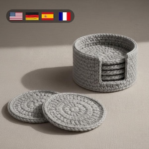 Puede incluir: Un juego de posavasos de ganchillo grises con un soporte a juego. Los posavasos son circulares, con un patrón en espiral. El soporte es cilíndrico con una ranura para guardar los posavasos. El juego está hecho de hilo suave y texturizado.
