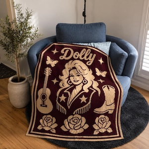 Puede incluir: Una manta de punto color granate y crema que presenta un retrato central de una mujer con cabello voluminoso, el nombre Dolly, una guitarra, una bota vaquera, mariposas y rosas. El diseño está enmarcado por un borde grueso a juego.