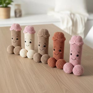 May include: A row of five handmade crochet penis dolls in various shades of brown and pink. Each doll has a smiling face and two crochet balls. The dolls are arranged on a light brown wooden surface.