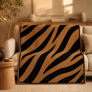 May include: A tan and black zebra-striped throw blanket on a beige sofa. The blanket has bold black stripes against a tan background. The design adds a touch of animal print to the room's decor. The blanket is a stylish home accessory.