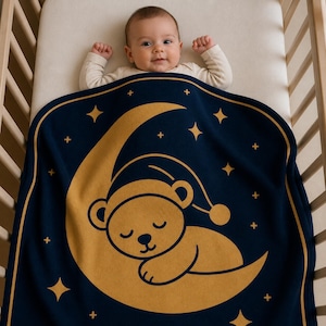 Op de afbeelding: Een baby in een wieg is bedekt met een marineblauwe deken met een gouden illustratie van een slapende beer op een maan, omringd door sterren. De baby draagt een witte romper.