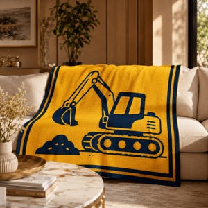 Peut inclure: Un plaid tricoté jaune orné d'un motif bleu foncé représentant une excavatrice de chantier creusant un tas de terre. La couverture est bordée de larges bandes bleu foncé sur les côtés gauche et droit, créant un design encadré.