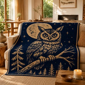 May include: A navy blue throw blanket with a detailed illustration of an owl perched on a branch, set against a full moon and starry sky. The owl and moon are in a tan colour, with tan trees at the bottom. The blanket is draped over a beige sofa.