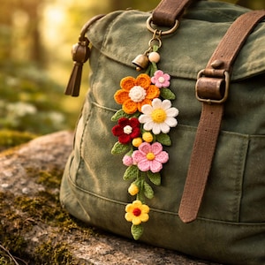 Peut inclure: Un charm fleuri au crochet aux couleurs vives, avec des fleurs orange, blanches, rouges, roses et jaunes, des feuilles vertes et une petite cloche. Le charm est accroché à un sac en toile verte avec des sangles en cuir marron.