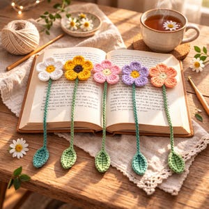 May include: An open book with six colorful crochet flower bookmarks. Each bookmark has a different color flower: white, yellow, pink, purple, and orange. The bookmarks have green stems and leaf-shaped ends. A cup of tea and a ball of yarn are also in the image.