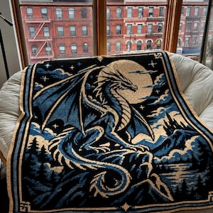 May include: A rectangular crochet blanket featuring a large dragon perched on a rocky cliff under a full moon. The design uses black, blue, and cream yarn to depict a night landscape with a castle, forest, and stars within a decorative border.