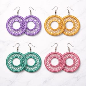 May include: Four pairs of hoop earrings in various colors: purple, gold, teal, and pink. Each pair features a circular design with a detailed, openwork pattern. The earrings are displayed on a white marble surface, showcasing their vibrant hues.